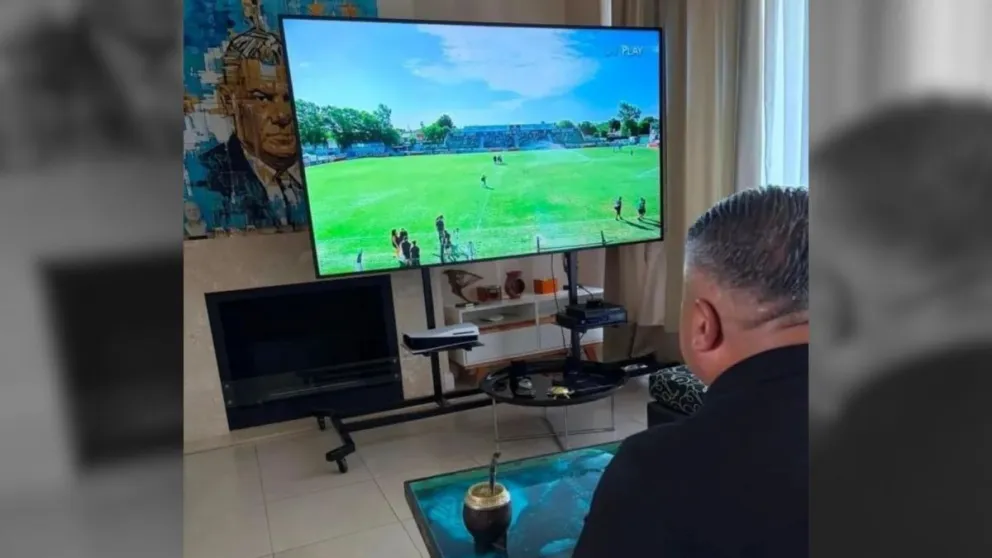 Tapia, con un retrato suyo de fondo en la parde, observando el partido San Miguel y Central Norte, por la Primera Nacional.